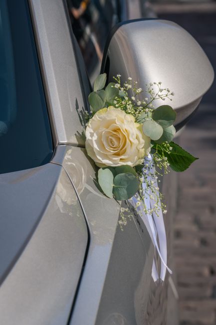A close-up of a white rose floral arrangement attached to the side mirror of a silver car, featuring a large, freshly bloomed white rose surrounded by eucalyptus leaves and delicate baby's breath. The