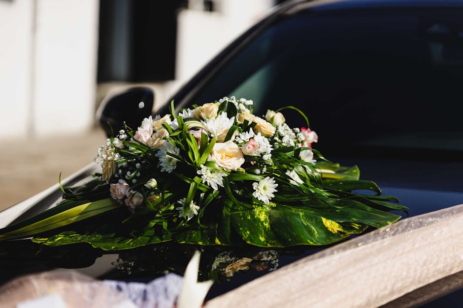 A close-up of a bridal bouquet arranged with white and pale pink roses, small white daisies, and baby's breath, accented with lush green foliage and long green leaves, resting on the hood of a sleek b