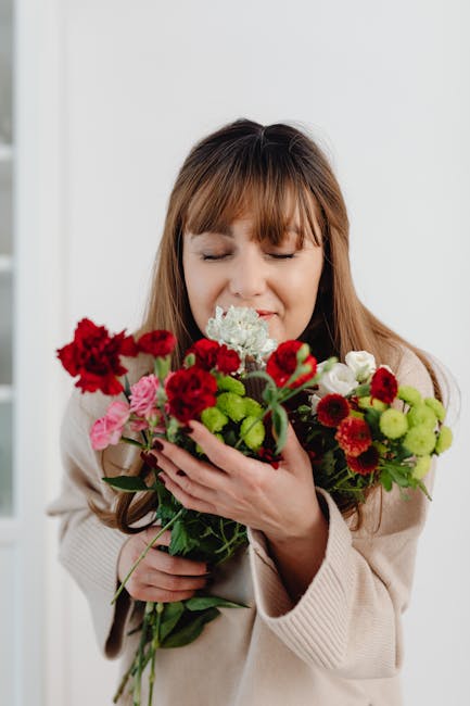 A woman with light brown hair and a beige sweater is smiling softly with her eyes closed while gently holding a bouquet of fresh flowers close to her face. The bouquet features vibrant red carnations,
