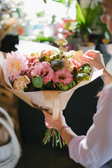 A person holding a large, vibrant floral bouquet wrapped in light brown paper with a cream-colored ribbon, featuring a mix of pink peonies, roses, and small white blossoms, accented with green foliage