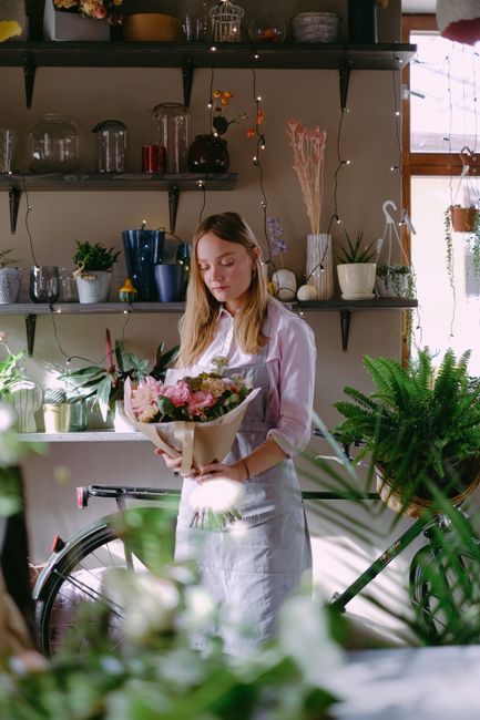 A young woman with long, light brown hair is standing indoors, holding a bouquet of fresh flowers wrapped in beige paper. The bouquet features pink and white blossoms, complemented by green foliage. S